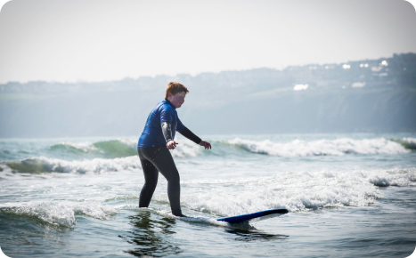 A male surfer in a blue wetsuit riding a dark surfboard on a small wave, with a foggy coastline and two tall structures visible in the distance.