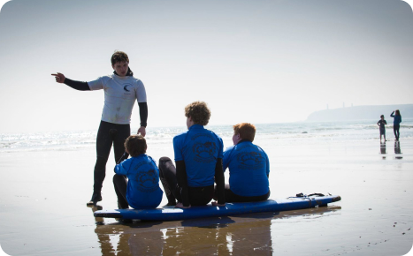 A surfing instructor in a white shirt and dark wetsuit bottoms pointing toward the ocean while giving a lesson to three young students sitting on a blue surfboard on a sunny beach.