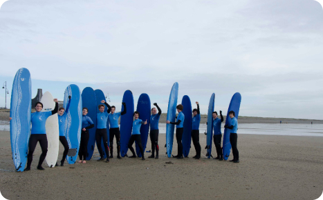 A group of male and female surfers in blue shirts and black wetsuits standing on a sandy beach with their blue and white surfboards.