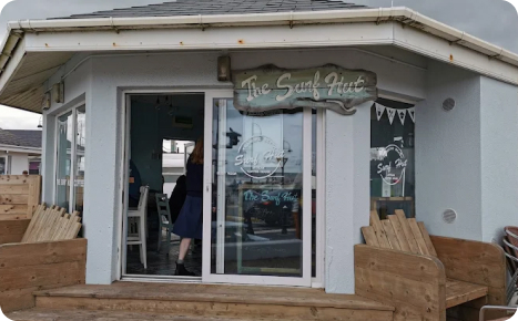 Exterior view of a small light gray building with a dark roof and large glass windows, featuring a wooden sign that reads "The Surf Hut" above the entrance.