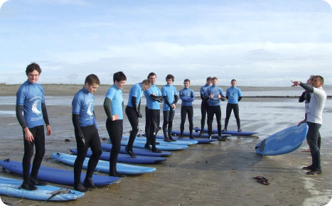 A large group of male surf students, wearing blue shirts and black wetsuit bottoms, practicing standing on blue surfboards laid out on a muddy beach, while an instructor points towards the sea.