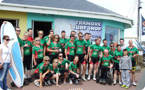 A large group of people in matching green and red cycling jerseys posing for a photo outside the "Tramore Surf Shop" on a sunny day.