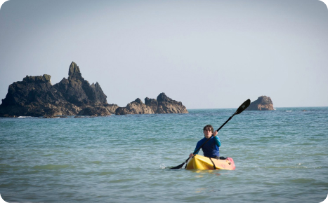 A person in a blue top paddling a yellow kayak in the ocean near a dramatic, sharp rock formation on a sunny day.