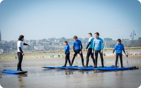 A group of people in wetsuits receiving surfing instruction on a sandy beach.