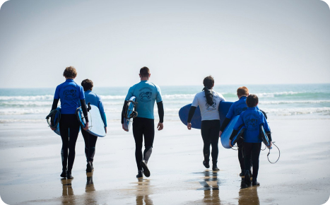 A group of six people, wearing wetsuits and carrying surfboards, walking away from the camera toward the ocean on a sunny beach.