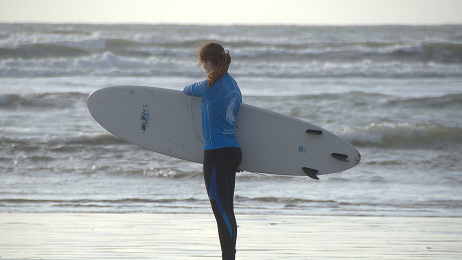 Female surfer in a blue shirt and black wetsuit bottoms standing on the beach, looking out at the waves while holding a white surfboard.