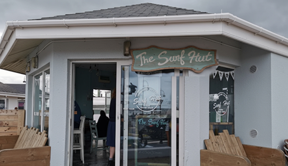 Exterior view of a small light gray building with a dark roof and large glass windows, featuring a wooden sign that reads "The Surf Hut" above the entrance.