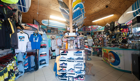Interior view of a brightly lit surf shop featuring a wide array of merchandise including clothing, shoes, flip-flops, bags, and multiple surfboards hanging from the wooden ceiling and displayed on racks.
