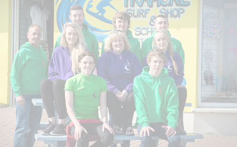 A group of nine staff members and instructors wearing green and purple hoodies and shirts posing outside the Tramore Surf Shop.