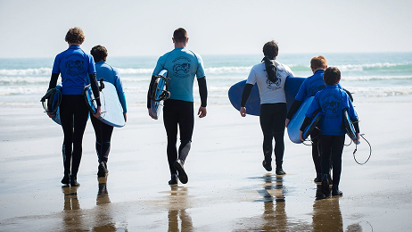 A group of six people, wearing wetsuits and carrying surfboards, walking away from the camera toward the ocean on a sunny beach.
